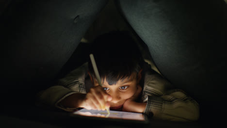Young-Boy-In-Home-Made-Camp-Made-From-Cushions-Playing-And-Drawing-With-Digital-Tablet-And-Stylus-At-Night-3